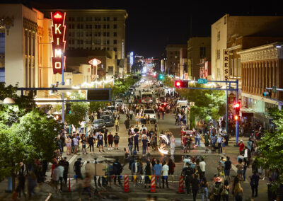 A bustling street night scene with people, featuring the KiMo Theater's neon sign and Kress building, capturing vibrant urban life and activity.