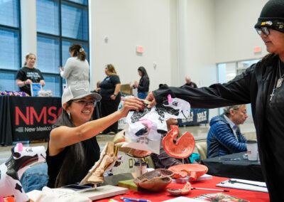 A person at a booth hands a model to another. Display includes anatomical models. Background features a banner with "New Mexico State."