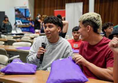 Group of people in a classroom setting, with a person holding a microphone and smiling. Participants are interacting and holding purple bags.