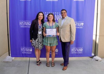 Three people stand in front of a blue Albuquerque Hispano Chamber of Commerce backdrop; the central person holds a certificate.