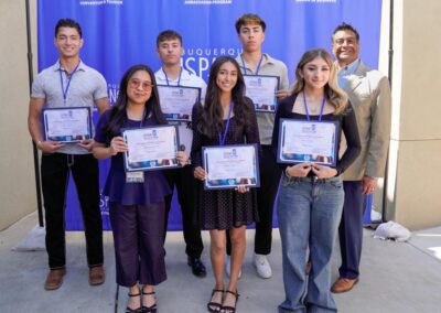 Seven individuals stand smiling with certificates in front of a blue background. They appear to be part of a recognition event.