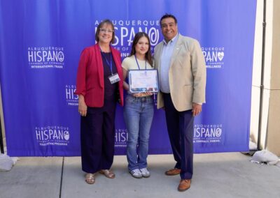 Three people stand in front of a blue banner displaying "Albuquerque Hispano Chamber of Commerce." One holds a certificate.