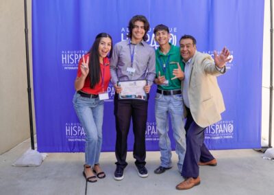 Four people pose cheerfully with a certificate in front of an "Albuquerque Hispano Chamber of Commerce" backdrop, depicting a celebratory event moment.