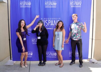 Four people pose in front of an Albuquerque Hispano Chamber of Commerce backdrop, smiling and gesturing energetically, wearing casual and semi-formal attire.