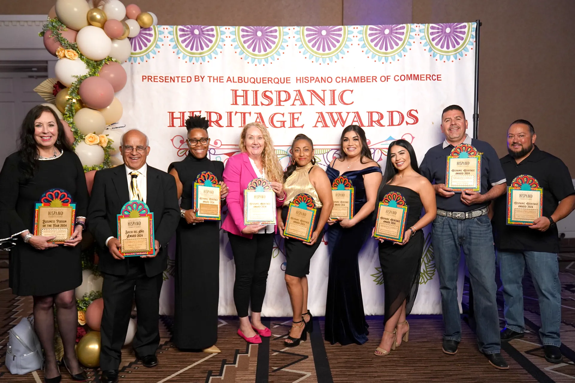 Nine people holding awards stand in front of a "Hispanic Heritage Awards" banner, decorated with balloons and flowers.