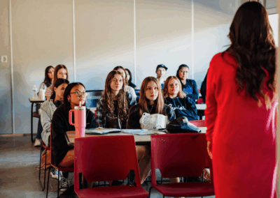 A person in red speaks to attentive students in a classroom setting with red chairs and desks, fostering an engaging learning environment.