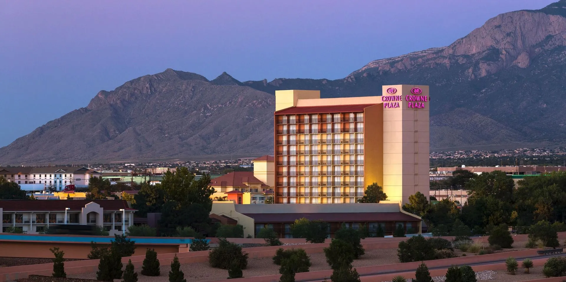 Crowne Plaza hotel against a backdrop of the Sandia Mountains, with surrounding trees and urban buildings under a clear purple-hued sky.